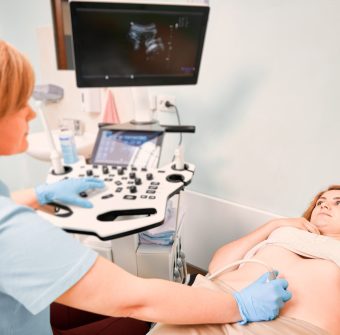 Young woman lying on daybed while doctor examining patient abdomen with ultrasound scanner. Sonographer performing ultrasound scanning. Concept of healthcare, sonography and ultrasound diagnostics.